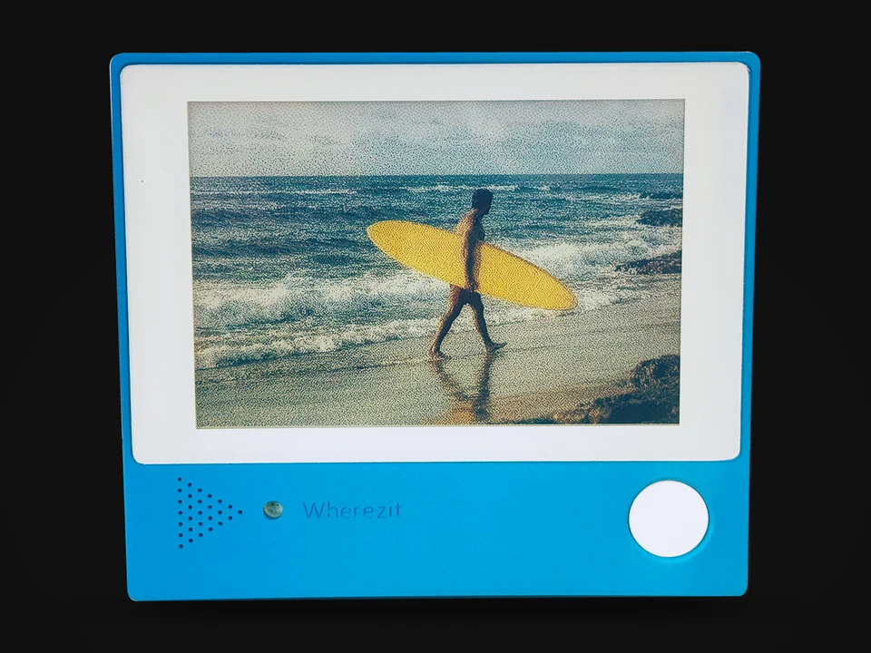 A blue Wherezit e-paper photo frame, a production unit on a black background, showing an example photo on its six-color screen.