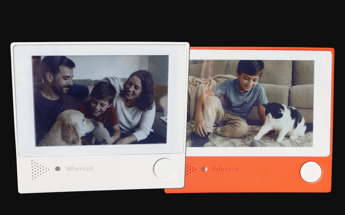Two Wherezit e-paper photo frames, production units in white and orange, on a black background, each showing an example photo on its screen.
